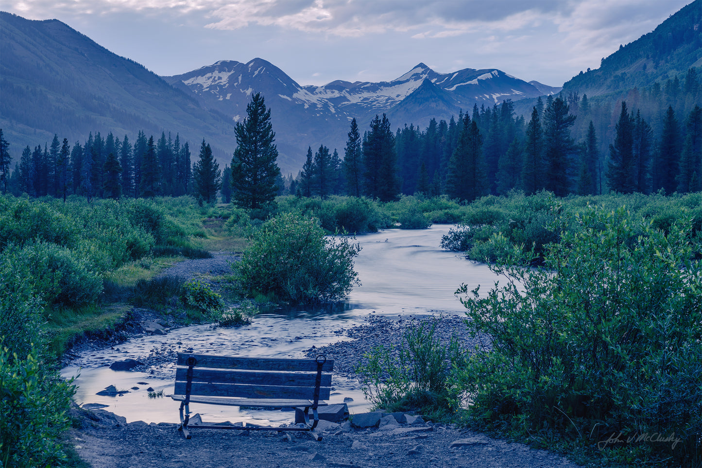 A peaceful dusk landscape image of a bench overlooking a gentle mountain river winding through willows and evergreens toward snowy mountains and a pink-hued sky.