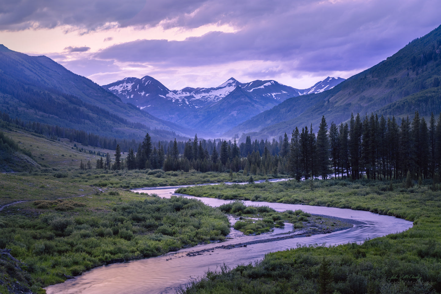 A winding river leads through a grove of evergreens to snowy mountains. The pale light of dusk illuminates the scene in this fine art landscape print by McClusky Nature Photography.