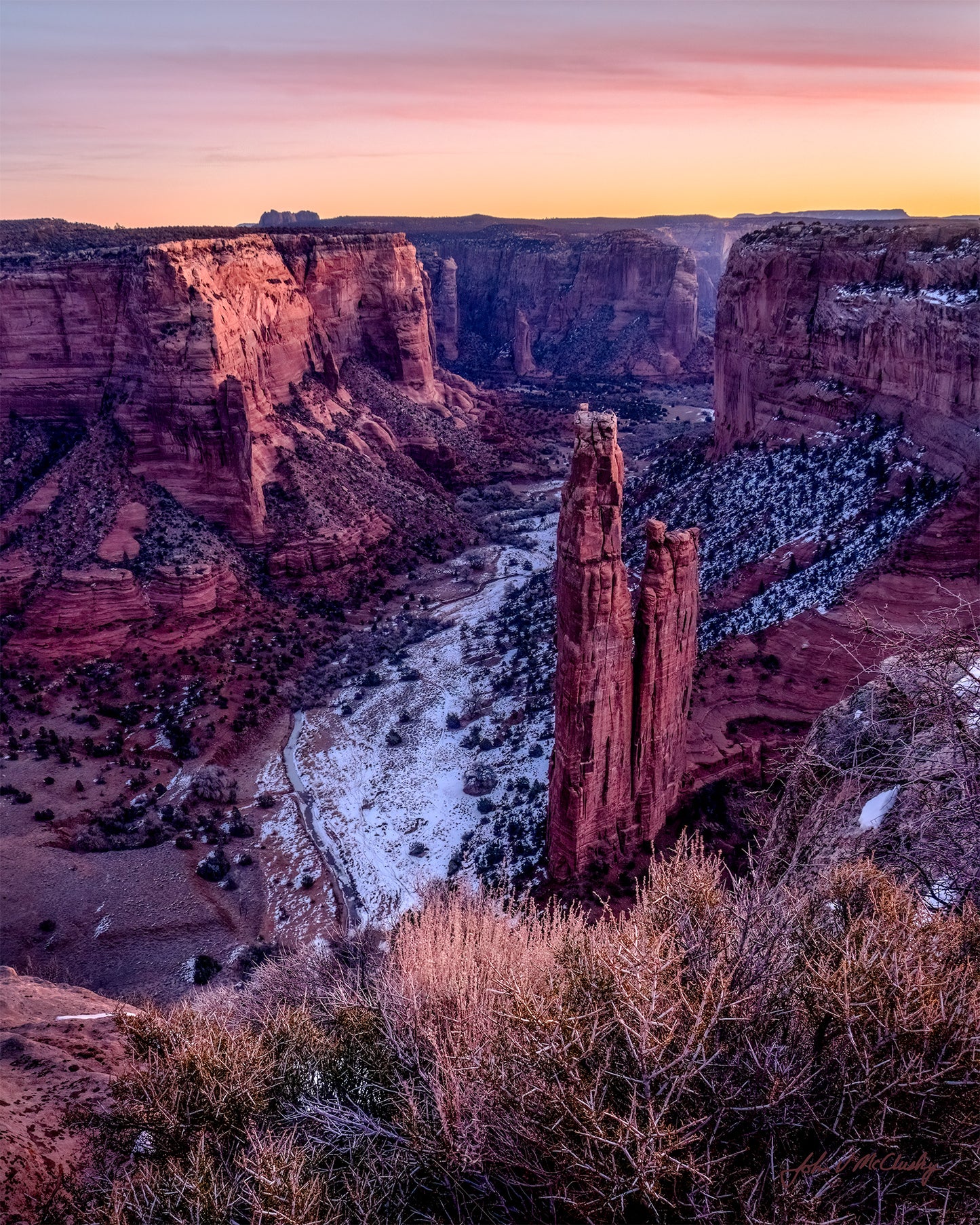 The pre-dawn glow lights the Canyon De Chelly. Wispy pink clouds grace the sky and the gentle light highlights the rugged canyon walls and the dusting of snow on the canyon floor.