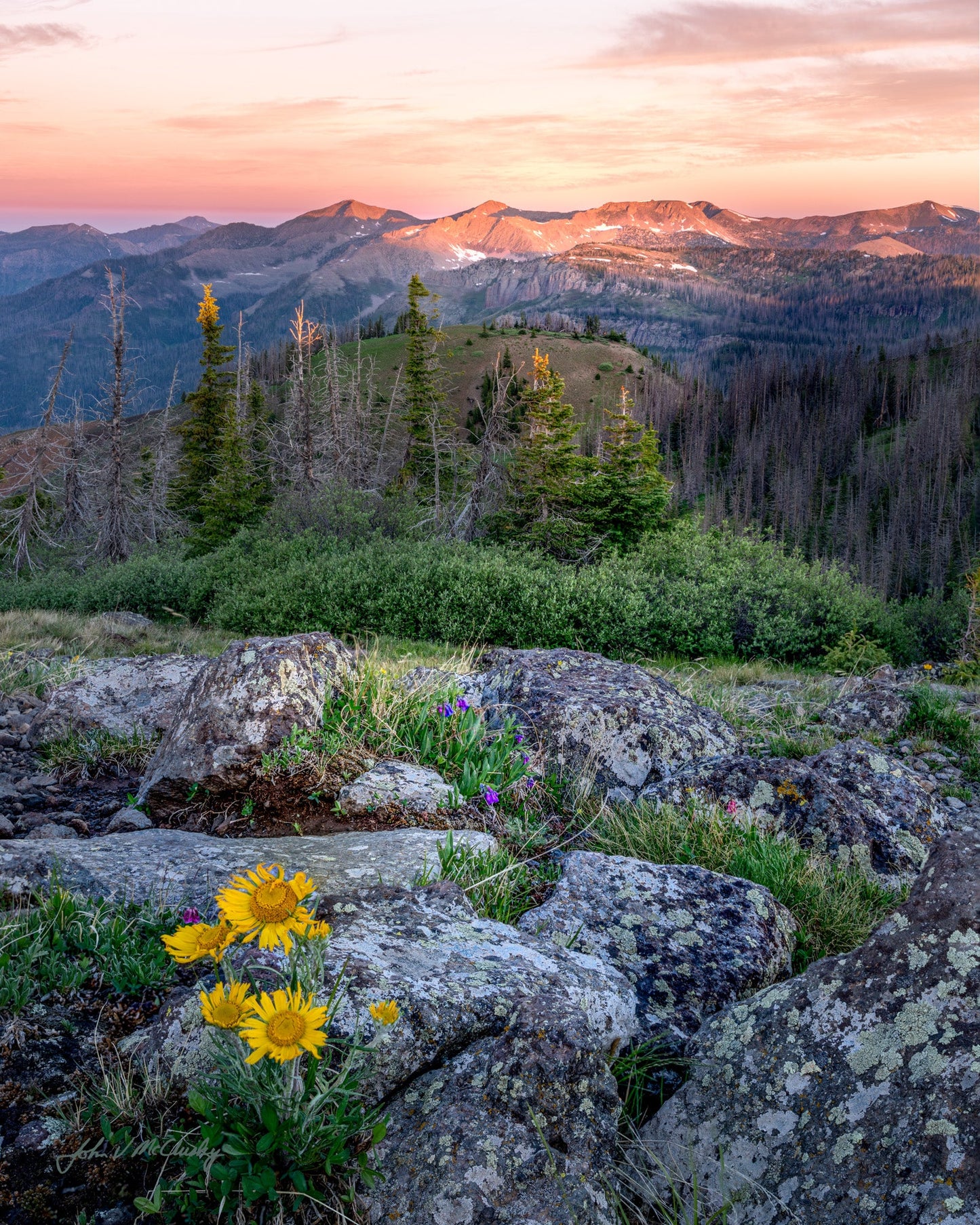 This image, titled "POSC Mountain Sunrise," showcases a breathtaking view of a mountain range at sunrise. The sky is painted in hues of pink and orange, contrasting beautifully with the rugged, gray rocks in the foreground.