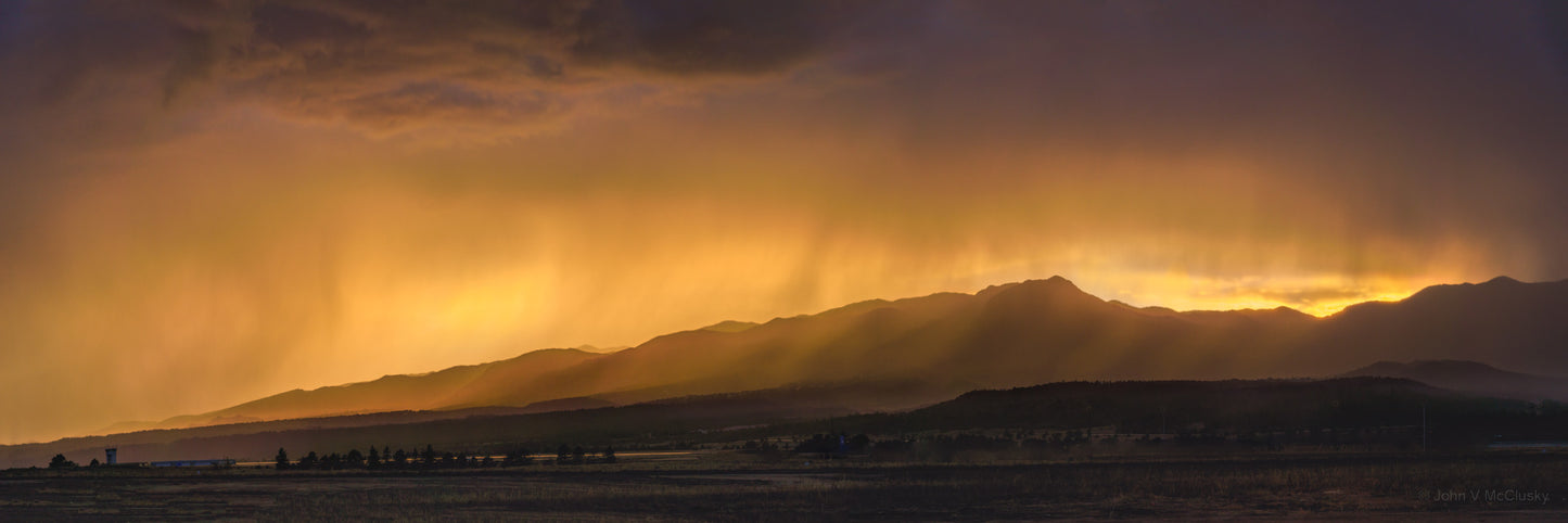 Clearing skies at sunset behind the Colorado Springs Front Range highlight streams of rain with a golden light. Panoramic landscape image by McClusky Nature Photography.