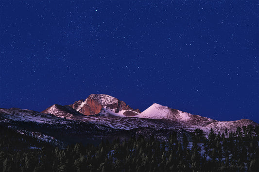 Stars shine above Long's Peak and the surrounding forest, lit with Alpenglow from the first light on the horizon.