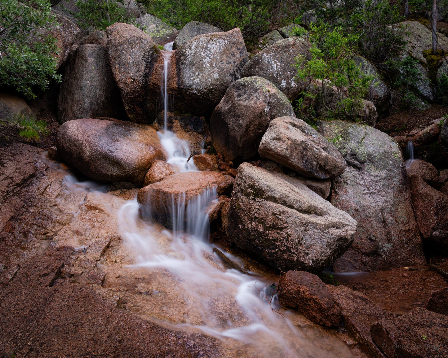 A waterfall cascades over granite rocks in the Pike National Forest. The early morning light emphasized the water and textured granite in this fine art print.
