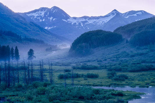a river flows through a misty valley in summer with snowy mountains above