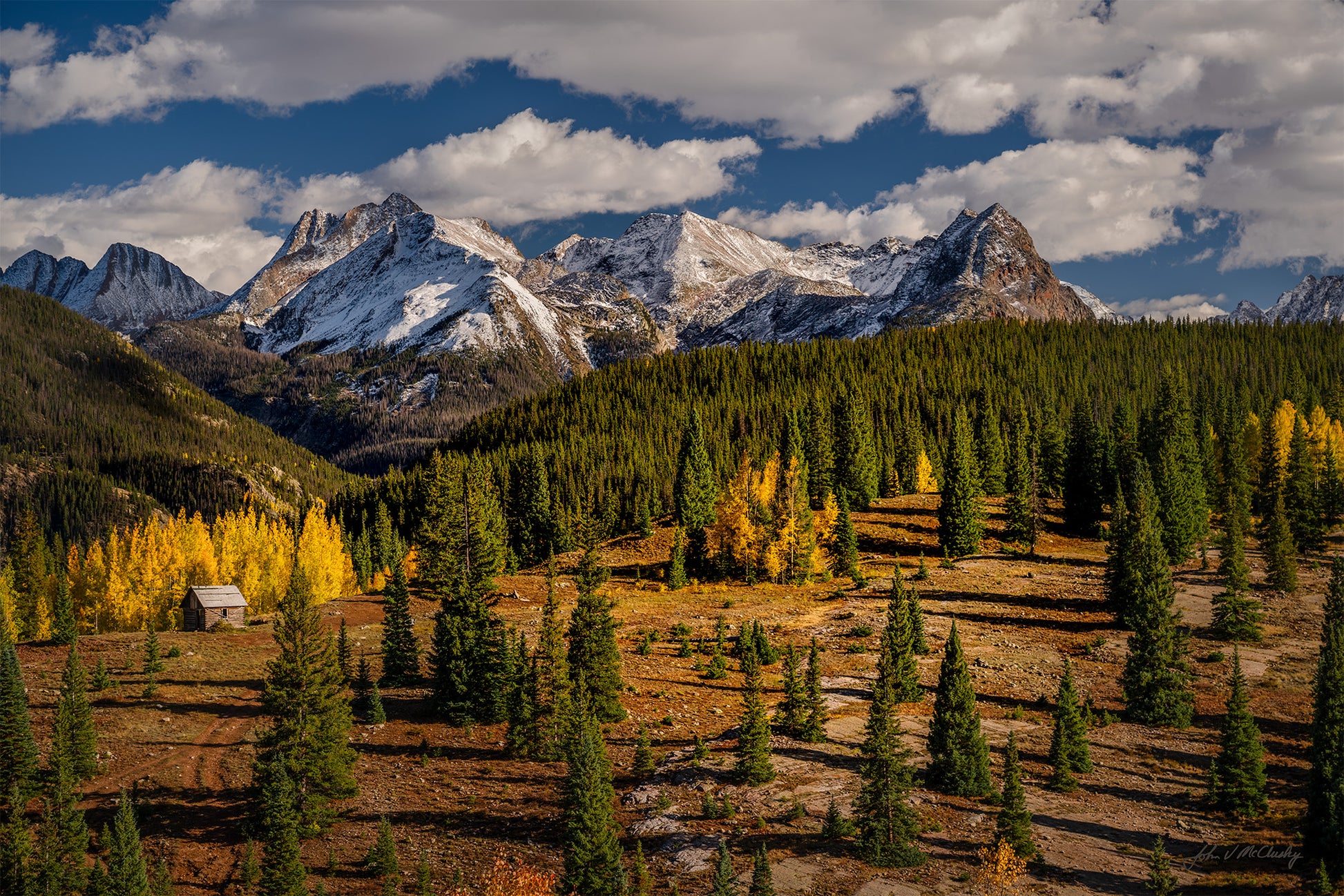 A lone cabin sits amid golden aspen with snow-clad San Juan mountains in the background