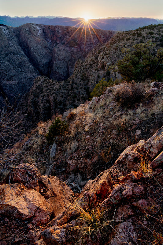 The rugged winter landscape of Colorado's Royal Gorge is emphasized by the last rays of the setting sun, highlighting multiple layers, from grasses amid the granite, to ridges withing the gorge, and finally to the distant mountains and shooting rays from the setting sun.