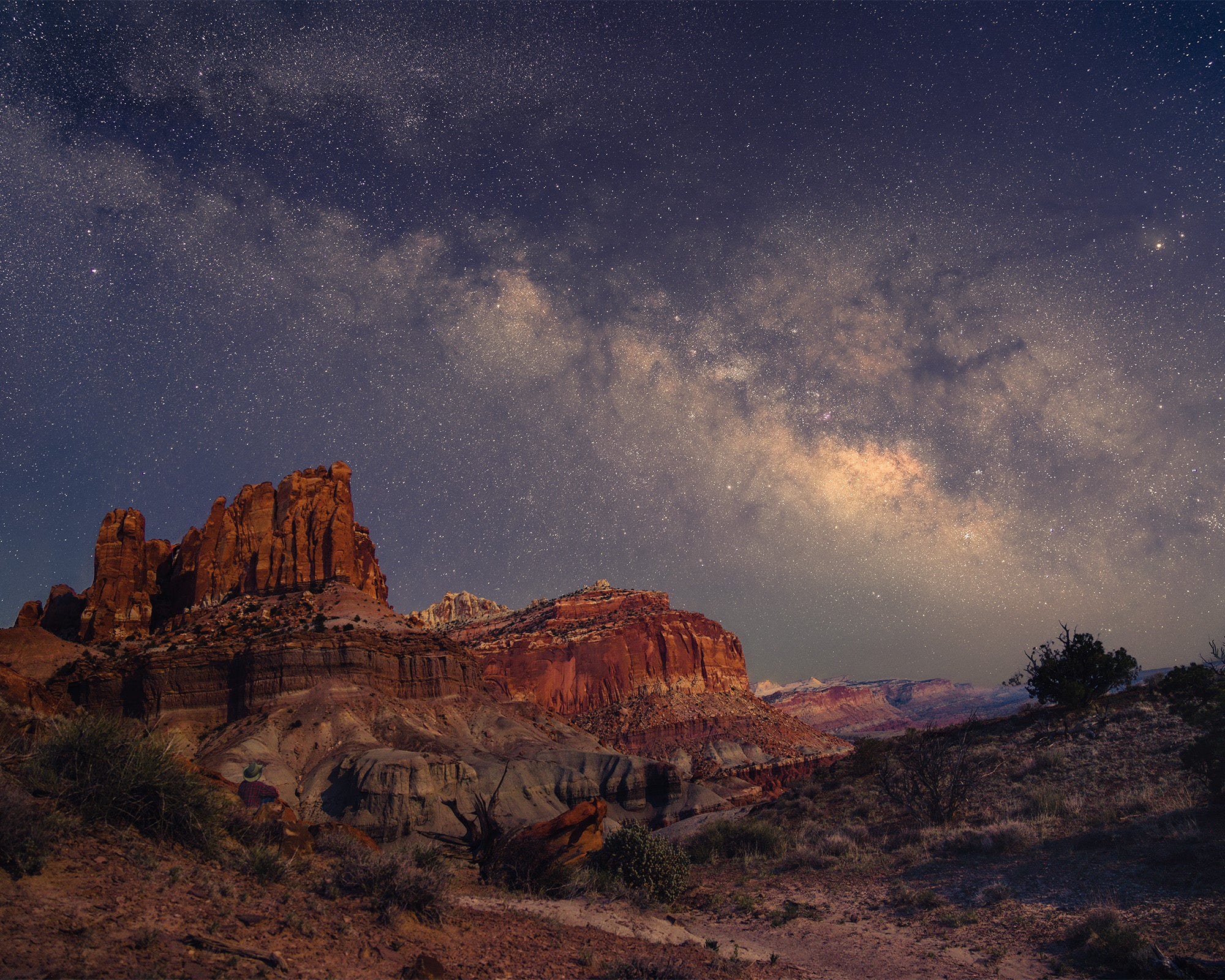 The Milky Way glows over the red cliffs of Capitol Reef National Park while a person sits and glories in the view.